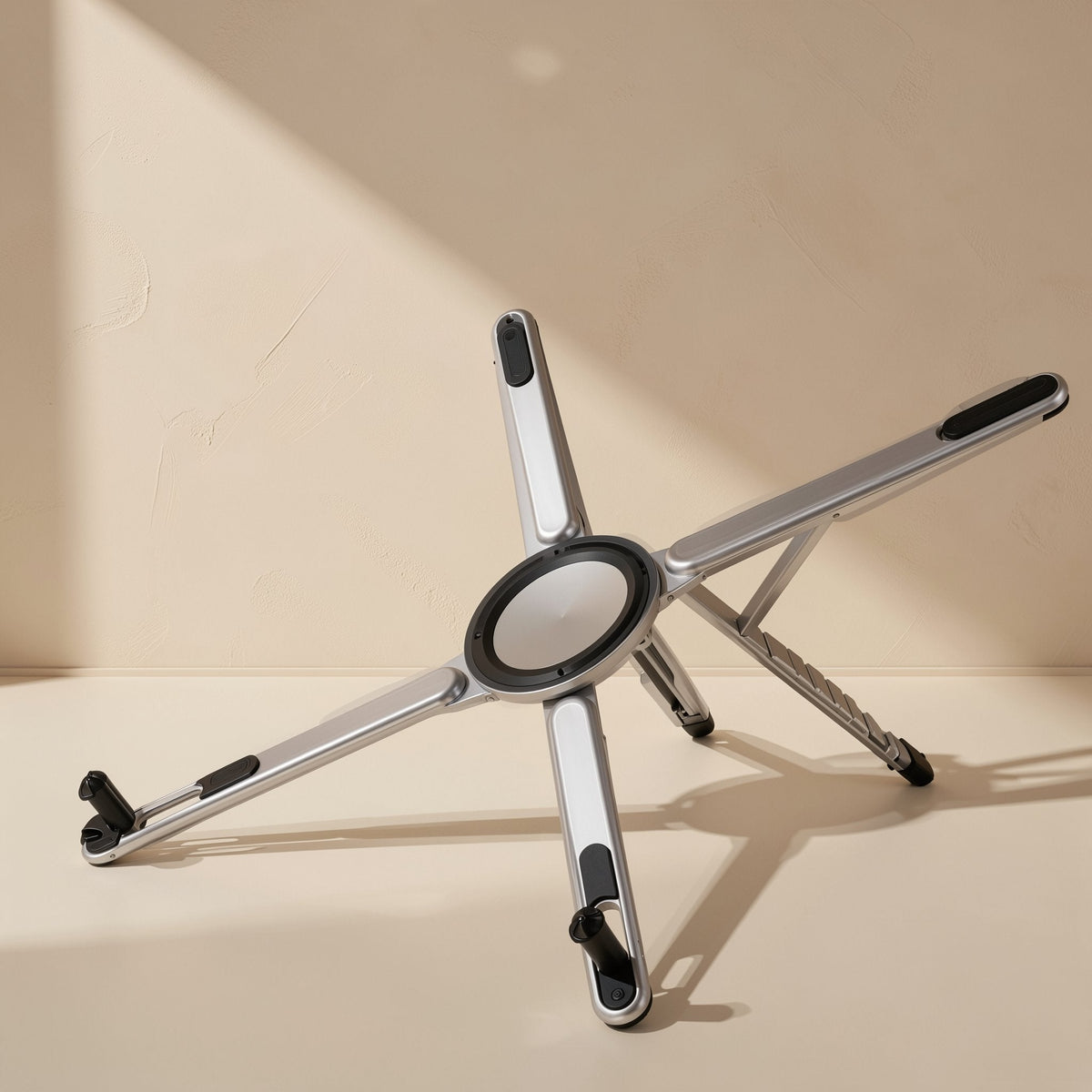 Alufold – Silver five-arm folding stand opened flat on a cream surface, showing the round center hub and black grips