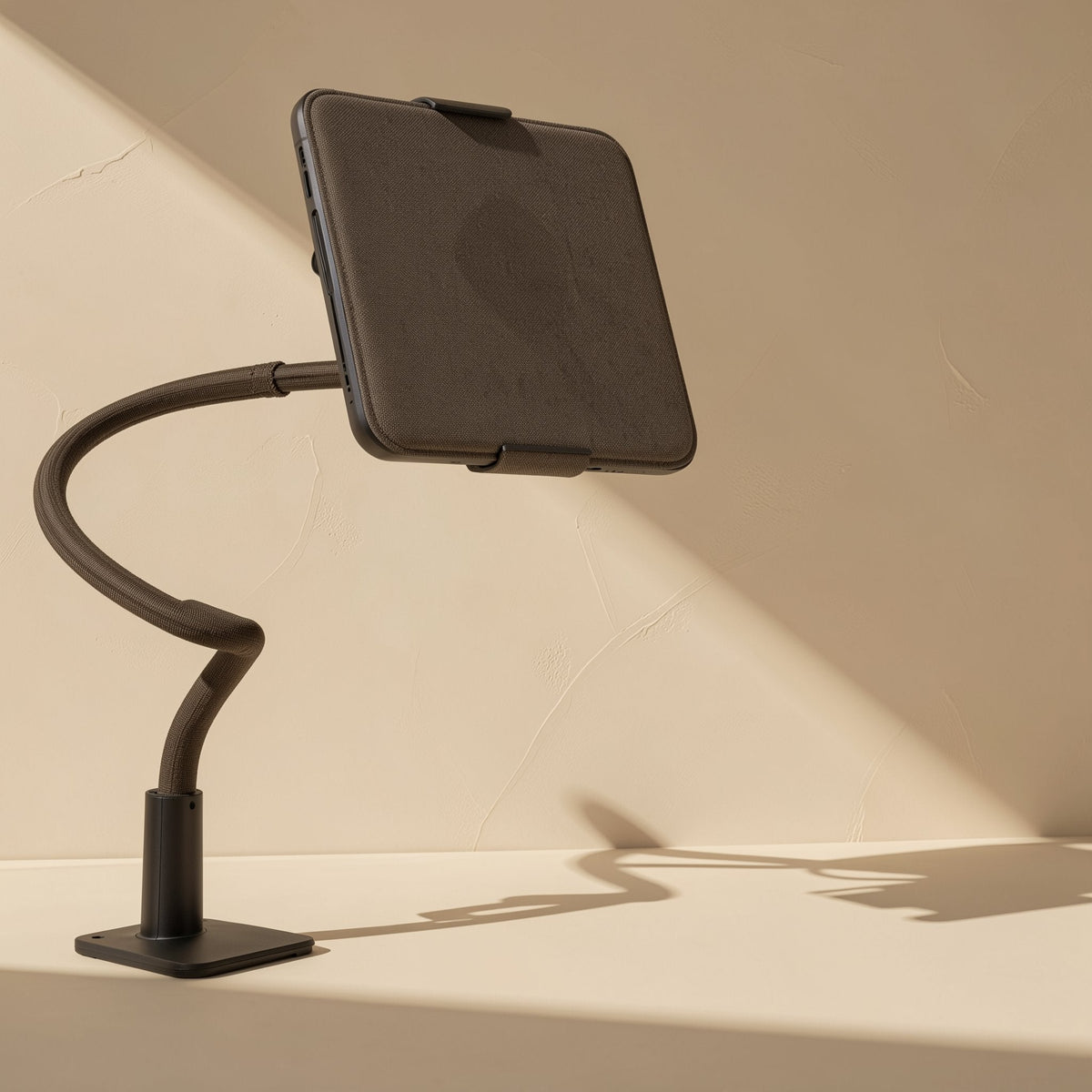 Rotaperch – Black gooseneck arm curving up from a square desk base, holding a tablet at a tilt in angled sunlight