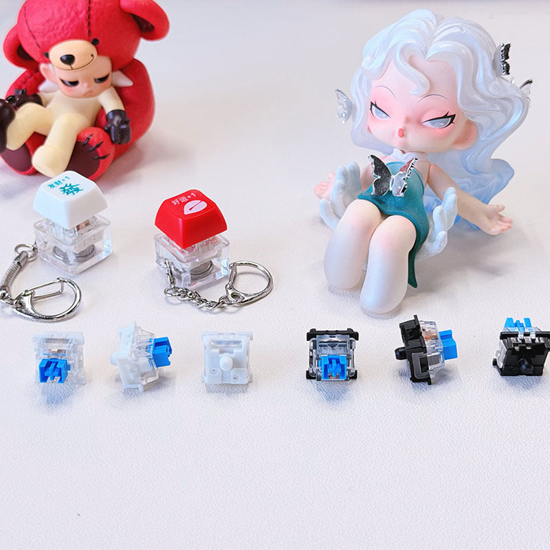 Crispop – Two keycap keychains above six loose blue and black mechanical switches, with a teddy figure and doll behind