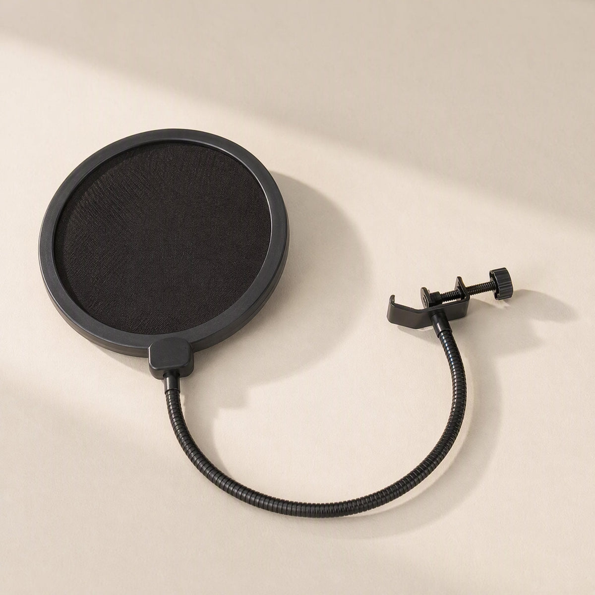 Streamveil – Black round pop filter with gooseneck and C-clamp lying on a beige surface in soft diagonal light