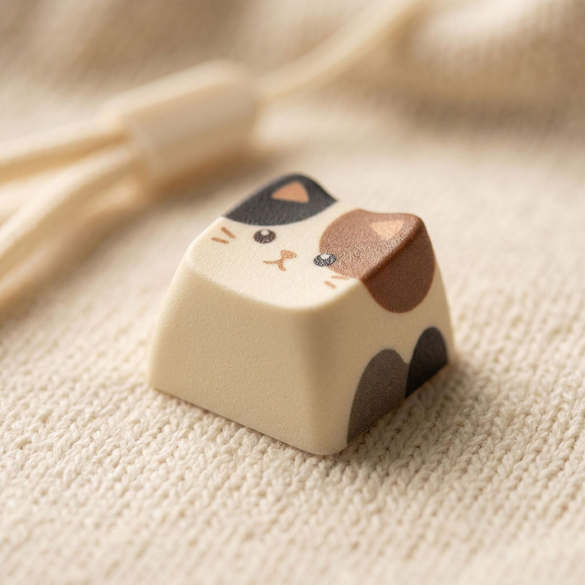Furrkey – Close-up of a calico cat keycap with black and brown patches on a cream knit fabric surface