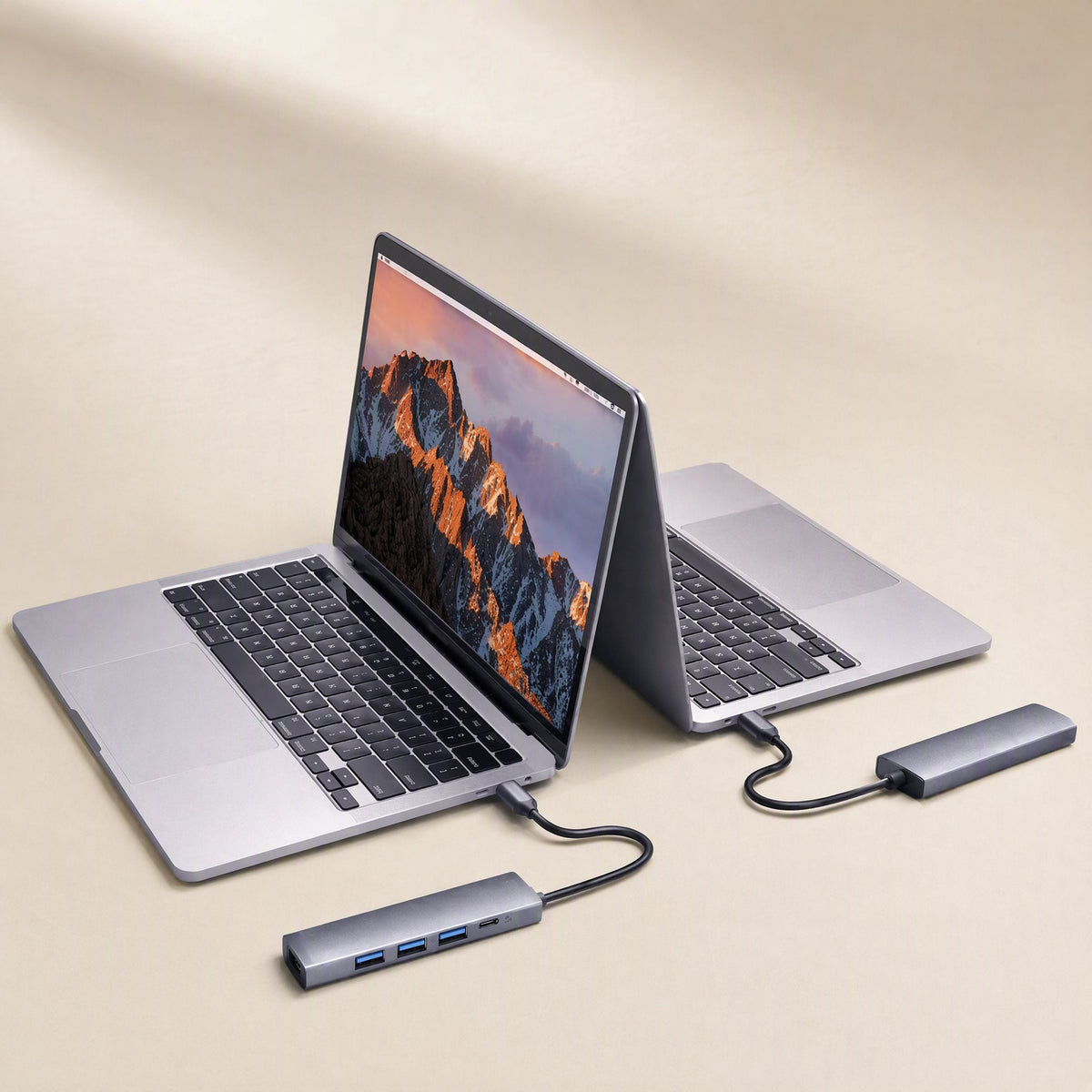 Dockbridge – Two laptops, one open and one half-closed, each linked to a slim gray USB-C hub on a beige surface