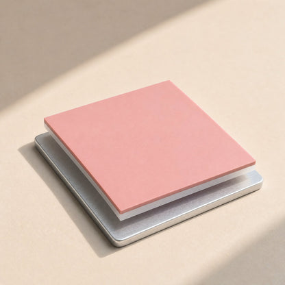 Thermaseal – Pink square thermal pad layered on a metal plate, shown at an angle on a beige surface