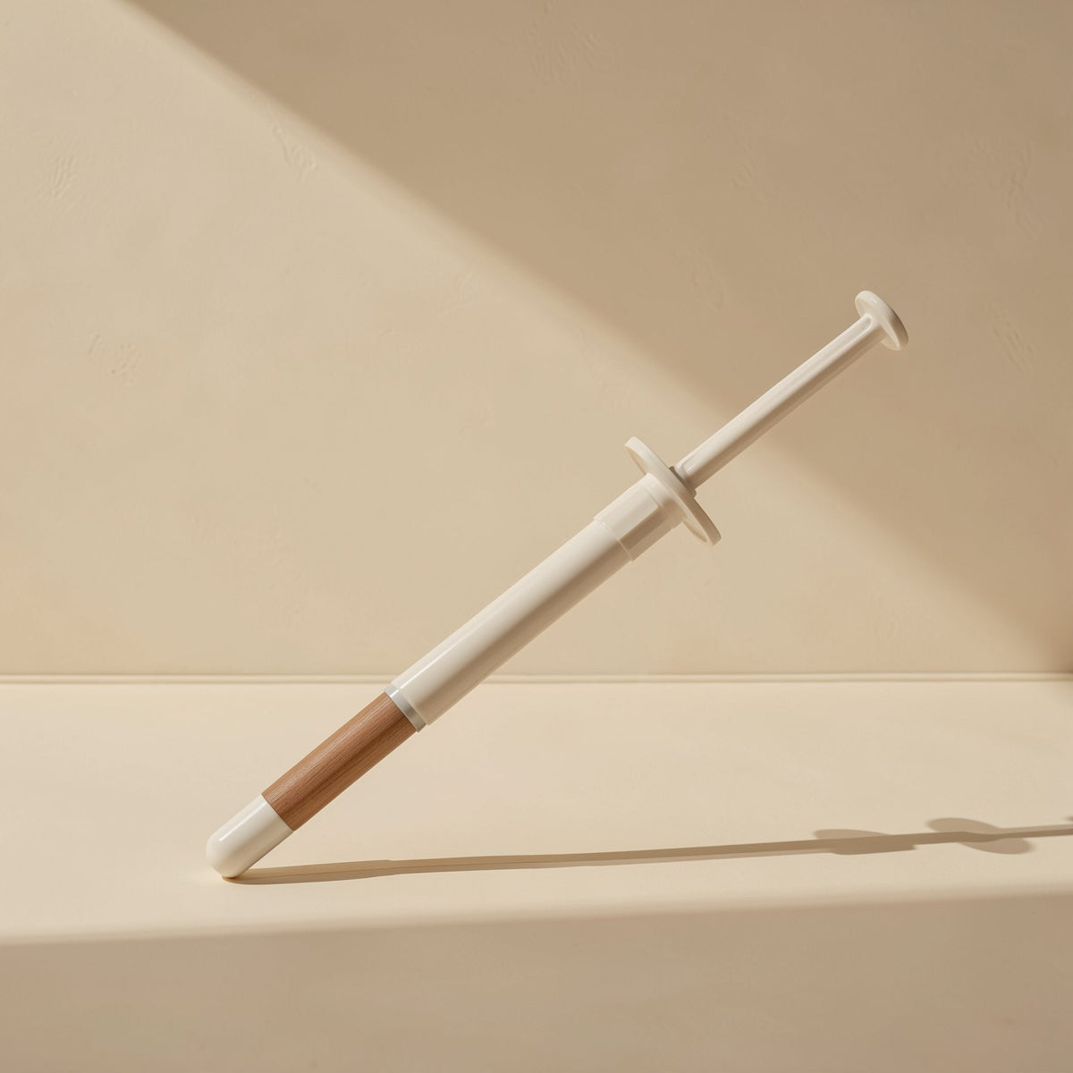 Heatpaste – Single syringe with white barrel and brown tip leaning at an angle against a beige wall in soft light