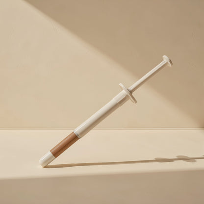 Heatpaste – Single syringe with white barrel and brown tip leaning at an angle against a beige wall in soft light