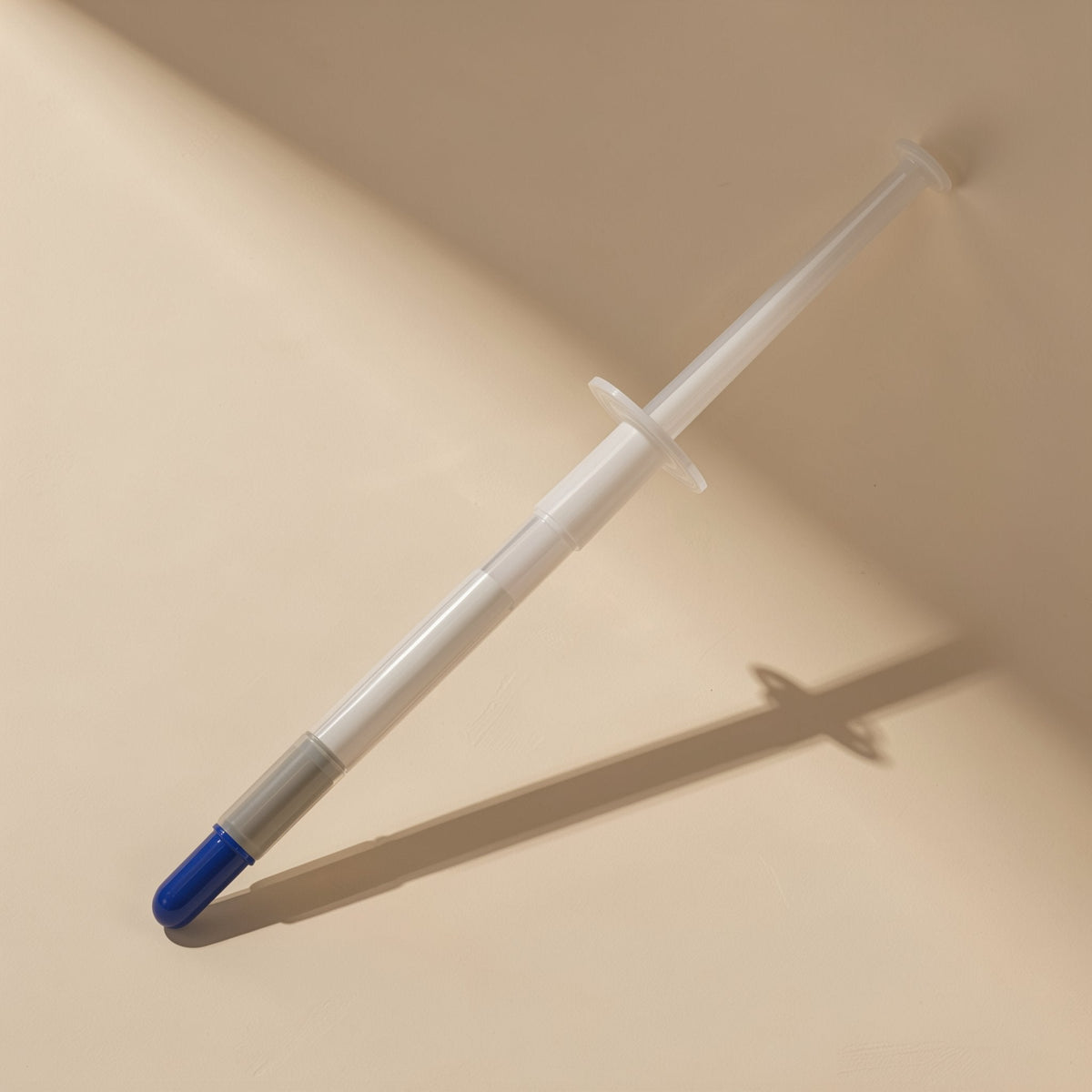 Heatpaste – Clear syringe with grey band and blue cap tip propped diagonally, casting a long shadow on beige