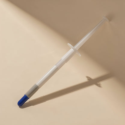Heatpaste – Clear syringe with grey band and blue cap tip propped diagonally, casting a long shadow on beige