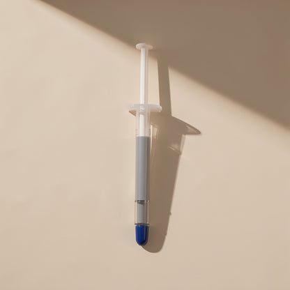Heatpaste – Upright syringe filled with grey paste, blue tip pointing down, against a cream wall with shadow