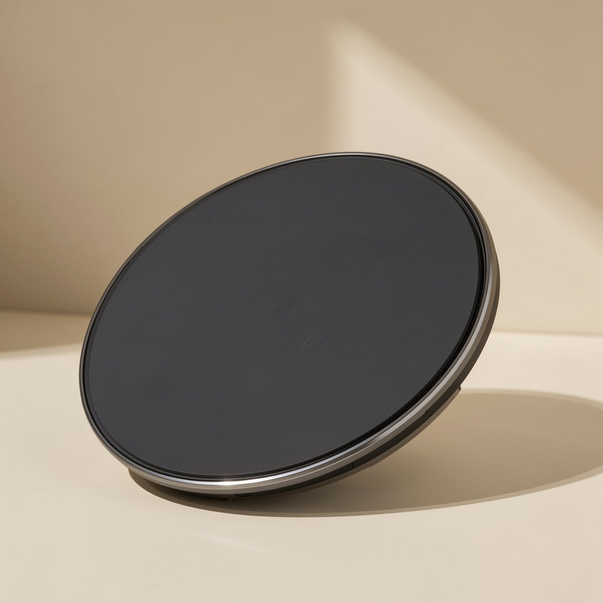 Voltflow Wireless Charger – black round charging pad with a thin metallic rim, tilted on a cream surface in angled sunlight