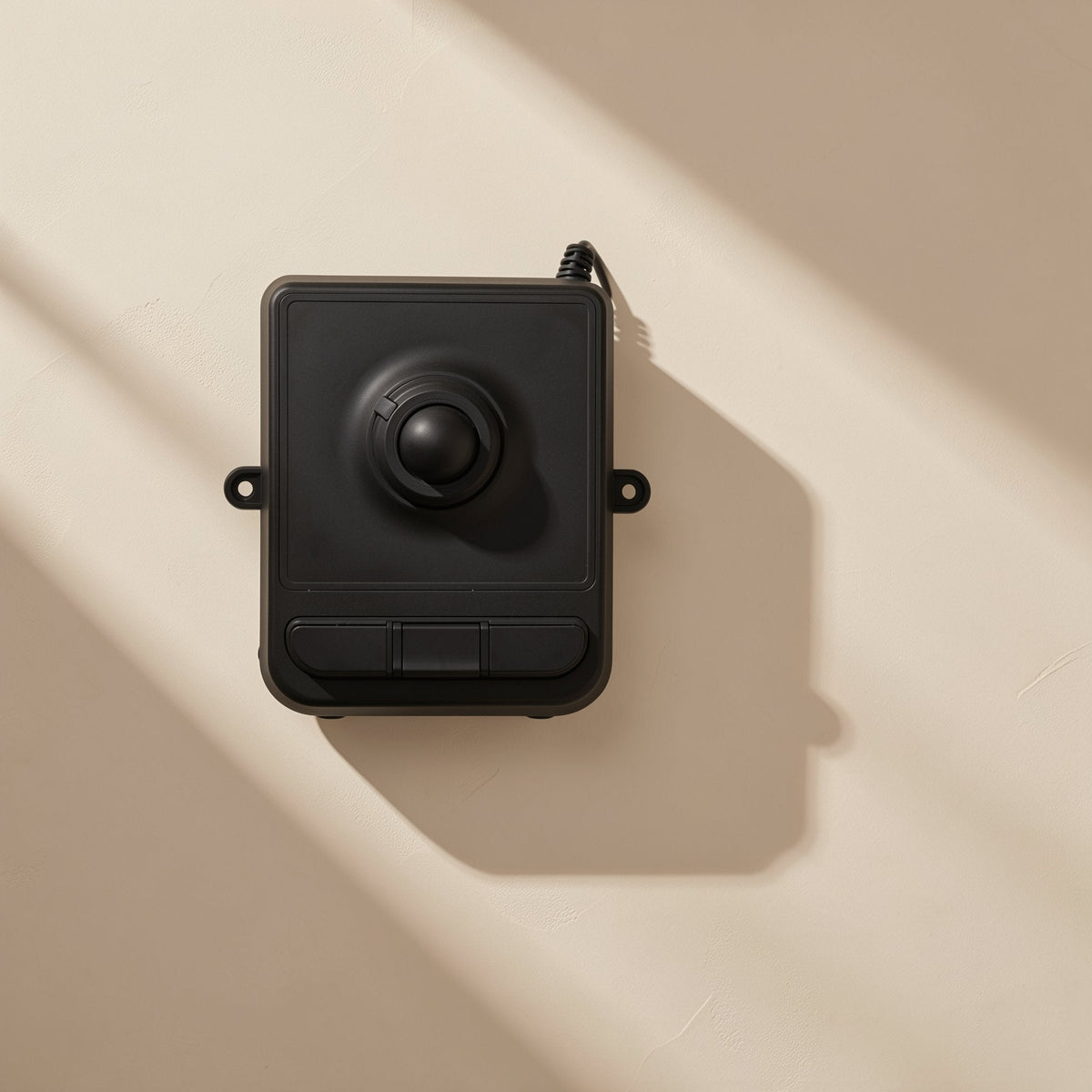 Trackroll – Black trackball mouse mounted on a beige wall, with cable at top and long diagonal shadows