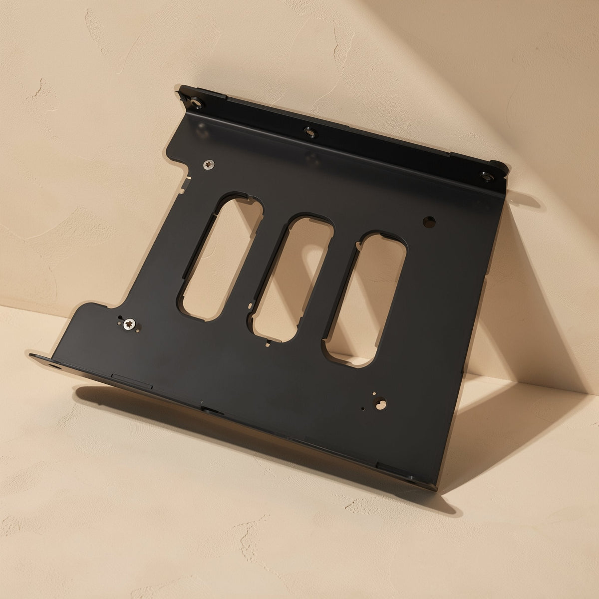 Diskcradle Desktop Hard Drive Bracket, black metal, propped at an angle against a beige wall in sunlight