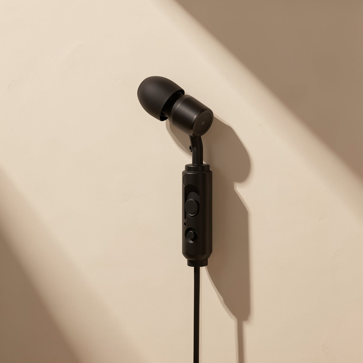 Cordtone – Earbud hanging from its black cable with inline remote and two buttons against a beige wall