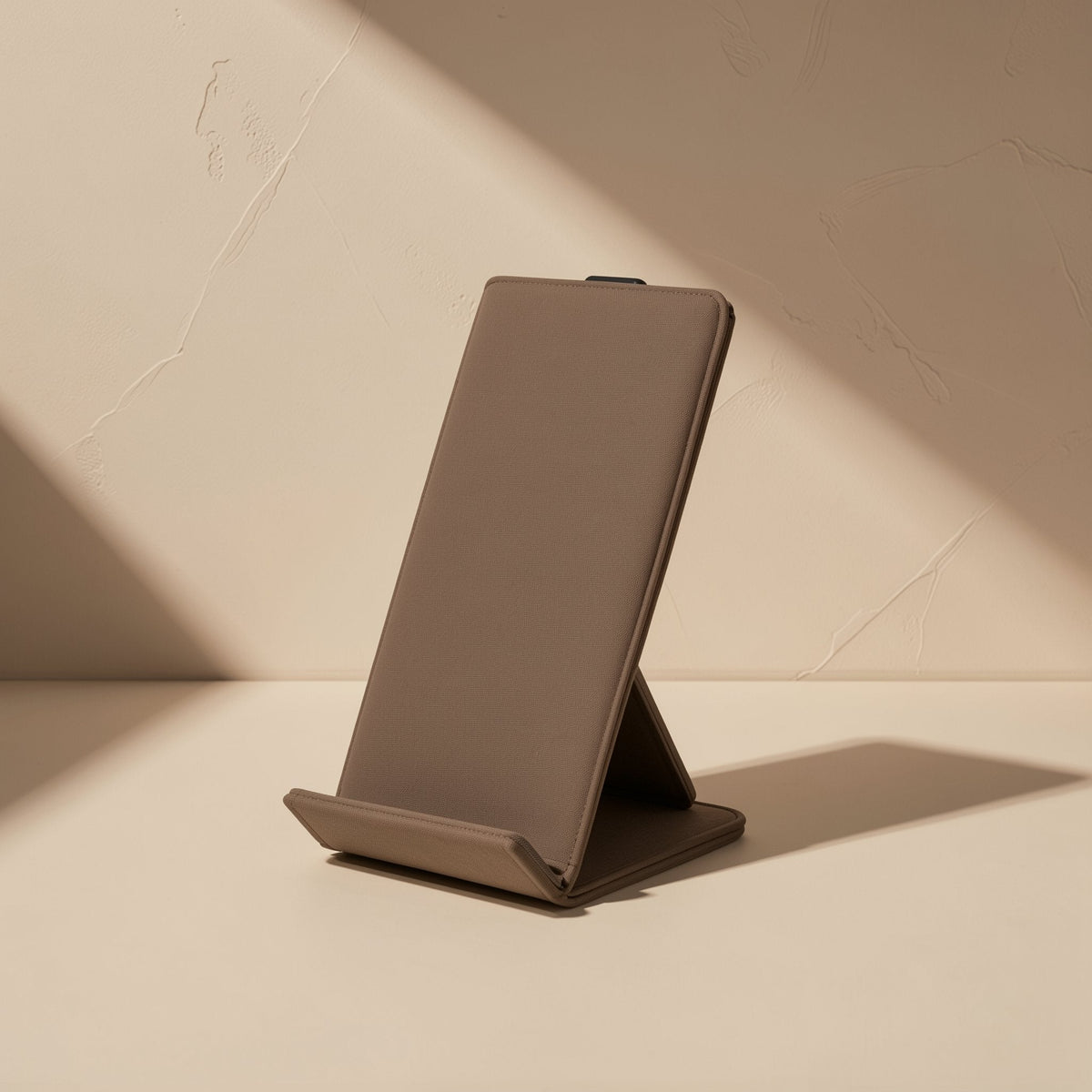 Tiltfold – Tan leather-look folding phone stand standing upright at an angle in warm sunlight against a plaster wall