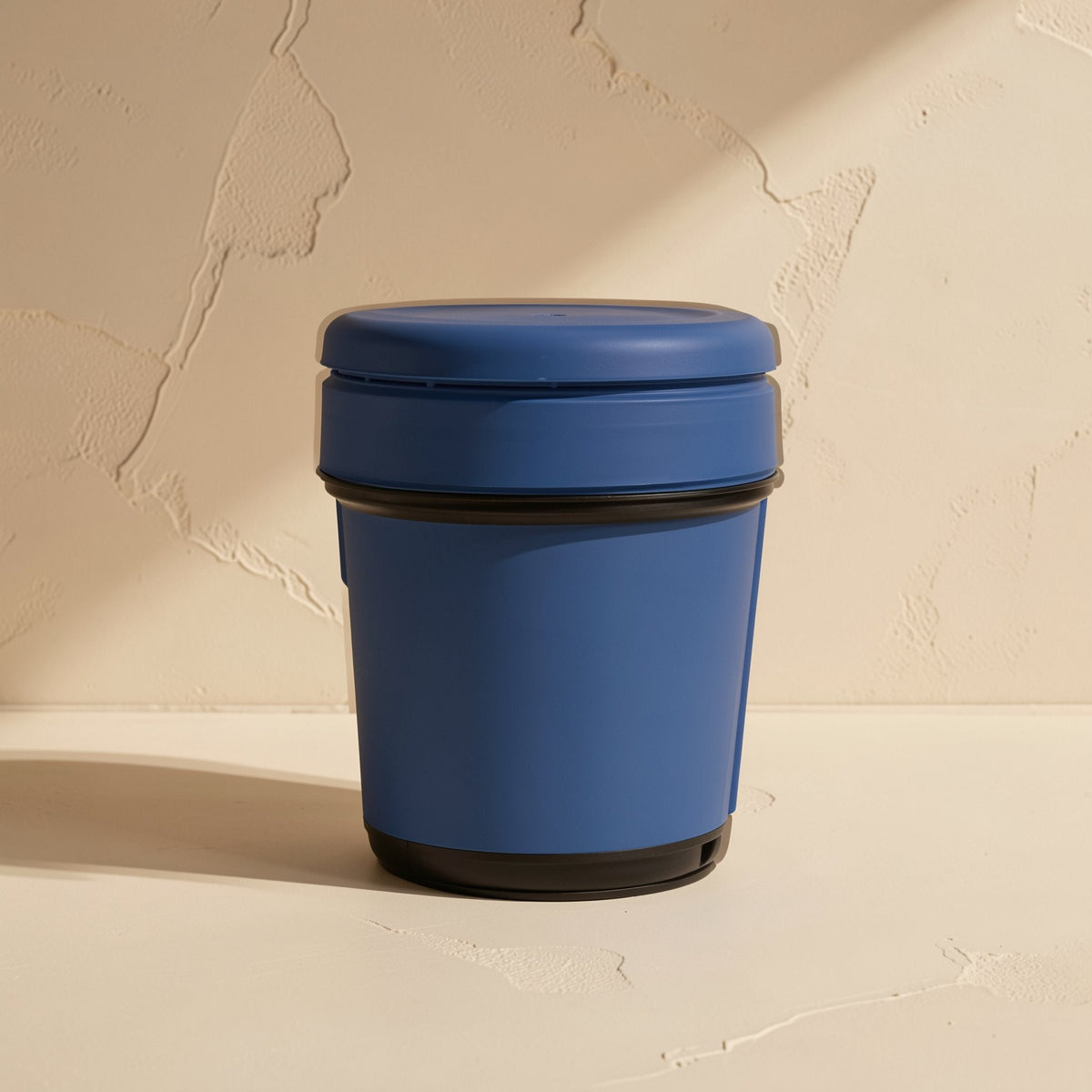 Pasteflow – Blue jar with black base and snap lid, side view against a textured beige wall in warm light