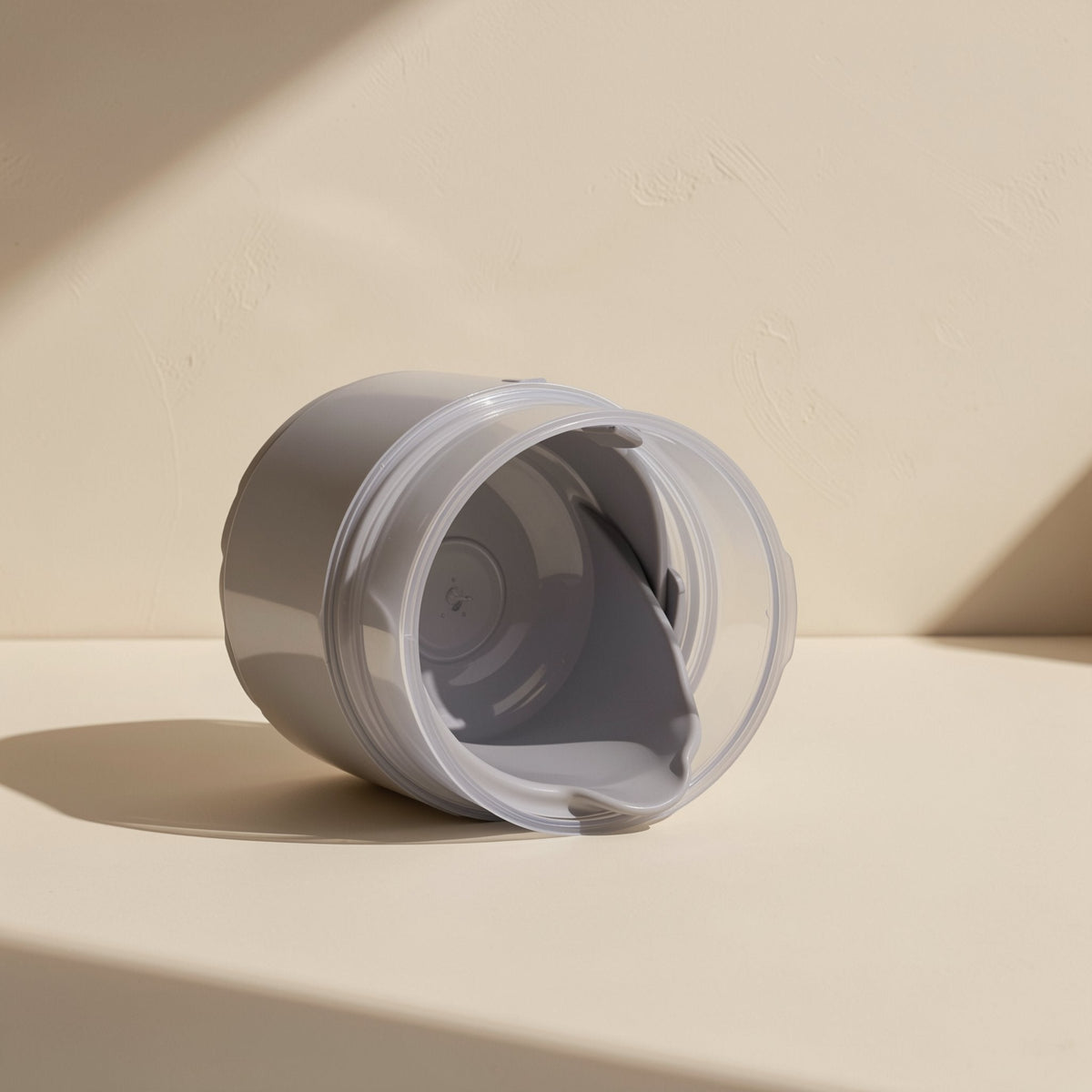 Pasteflow – Gray jar lying on its side with the clear inner lid open, showing the hollow interior