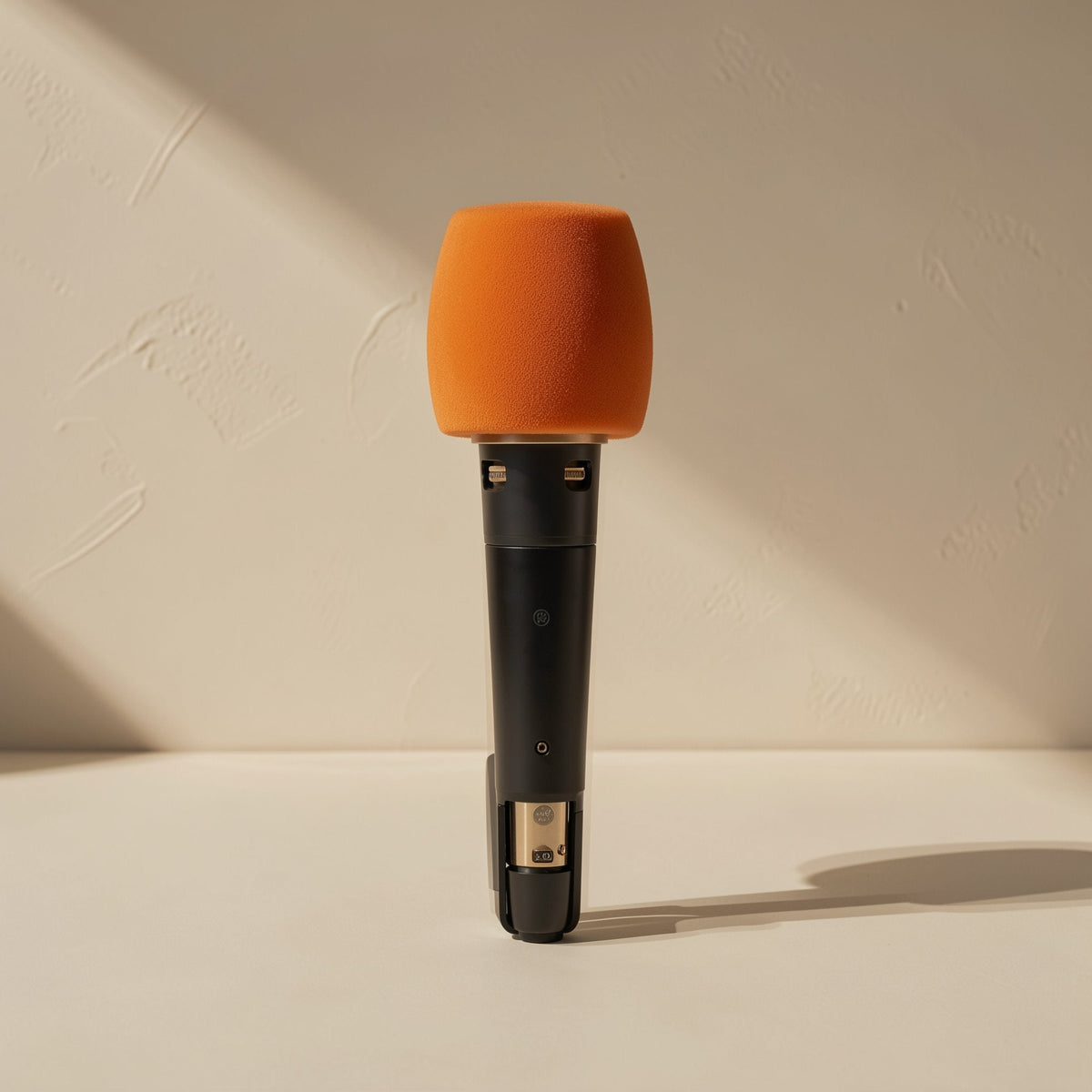 Micmuffle – Orange foam microphone cover fitted on a black wired handheld microphone, standing against a beige wall