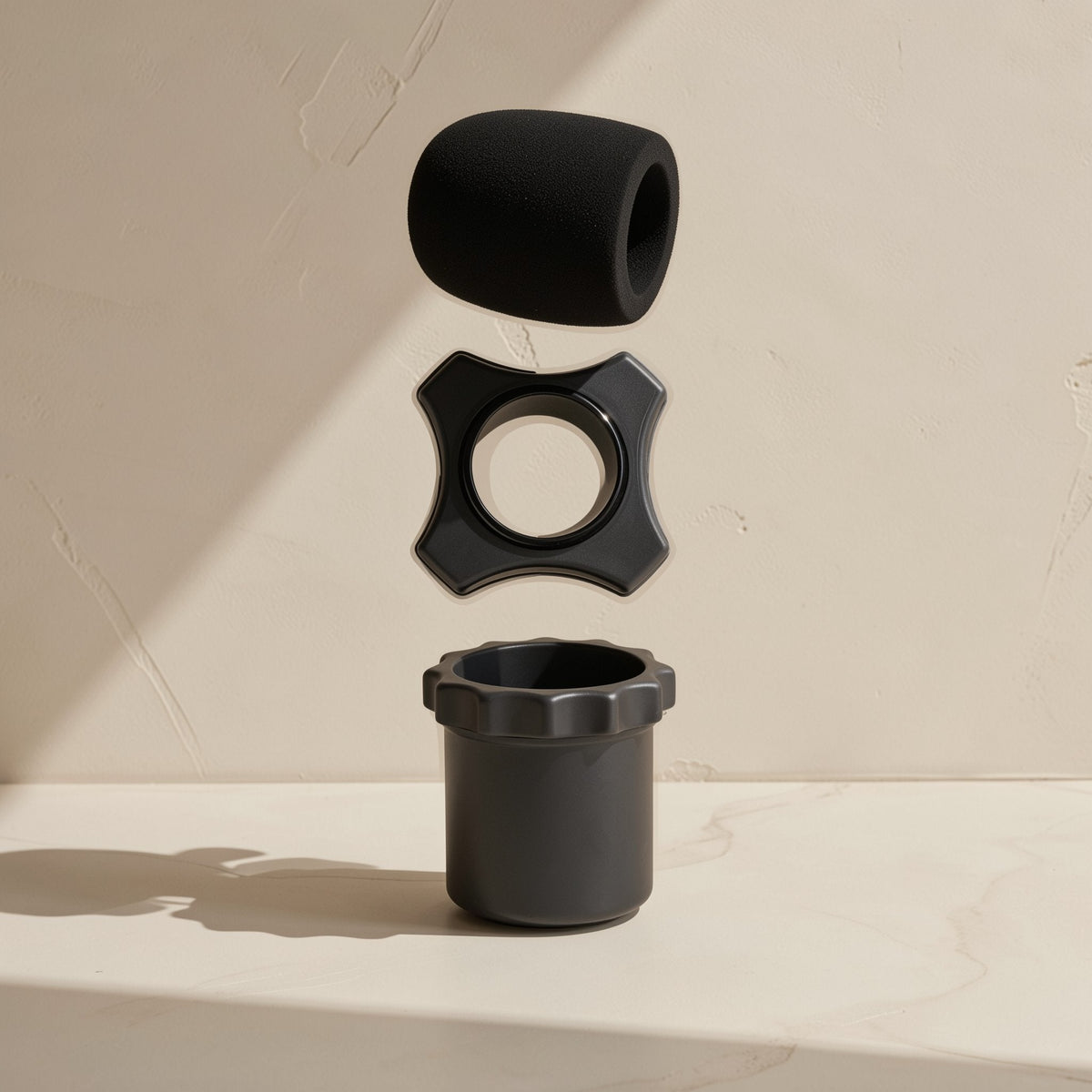 Micmuffle – Black foam microphone cover shown above a black four-point ring and cylindrical base in an exploded view