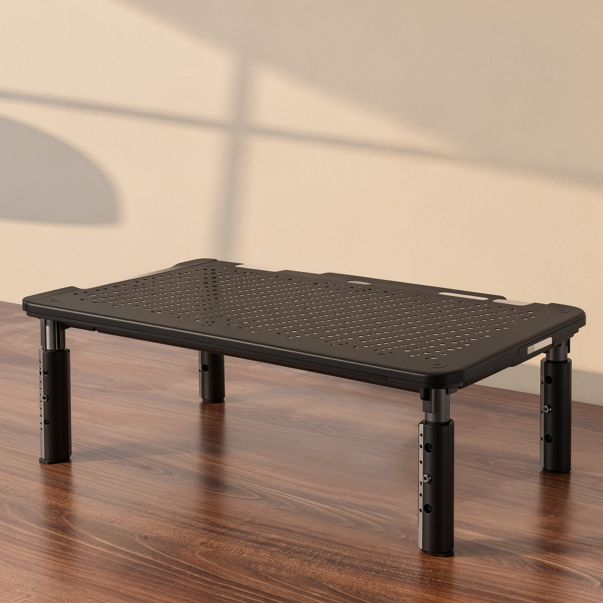 Screenlift – Black adjustable-leg stand with perforated top on a wood desk, seen from a low angle against a cream wall