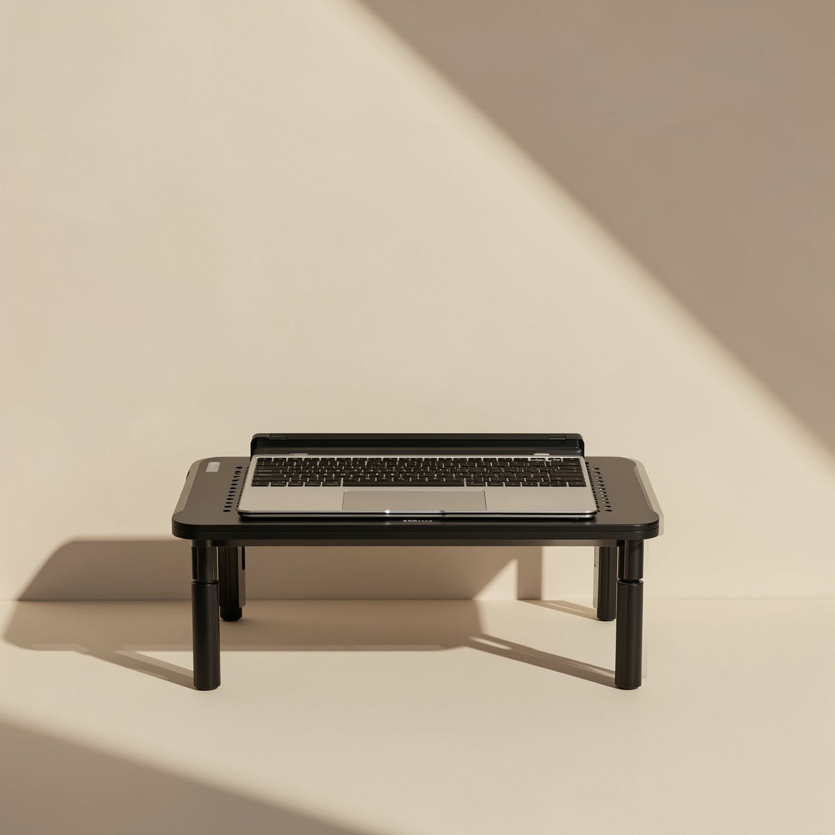 Screenlift – Laptop resting on the black stand, shown from the front with sunlight and shadow on a plain cream wall