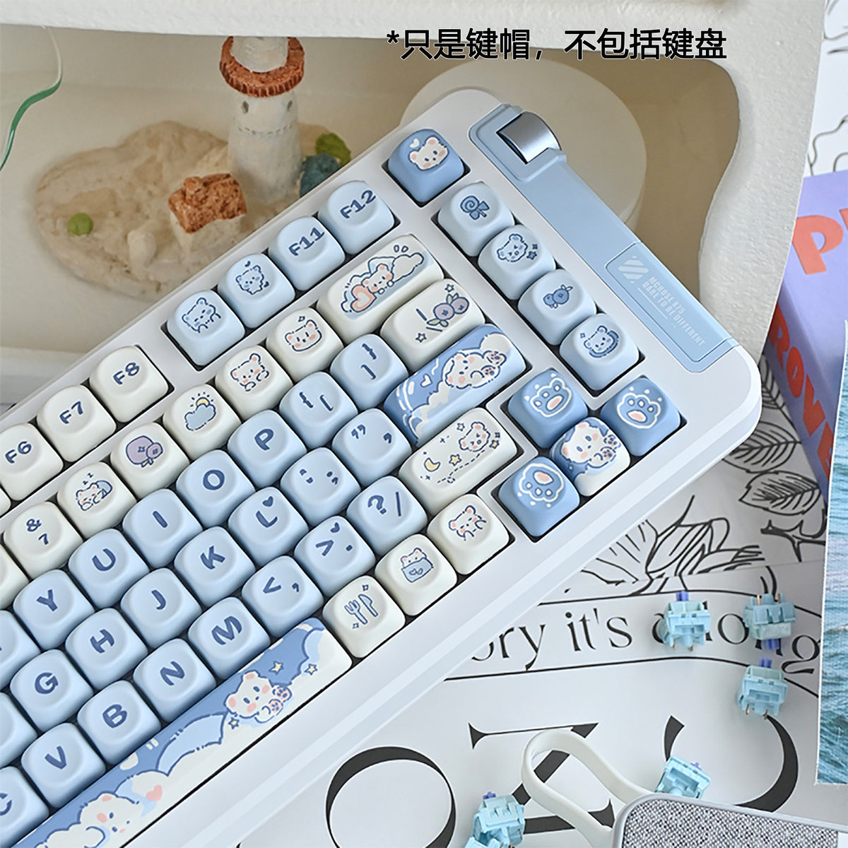 Cubicle Ice Cream Bear keycaps in blue and cream on a white keyboard, angled view beside a small lighthouse