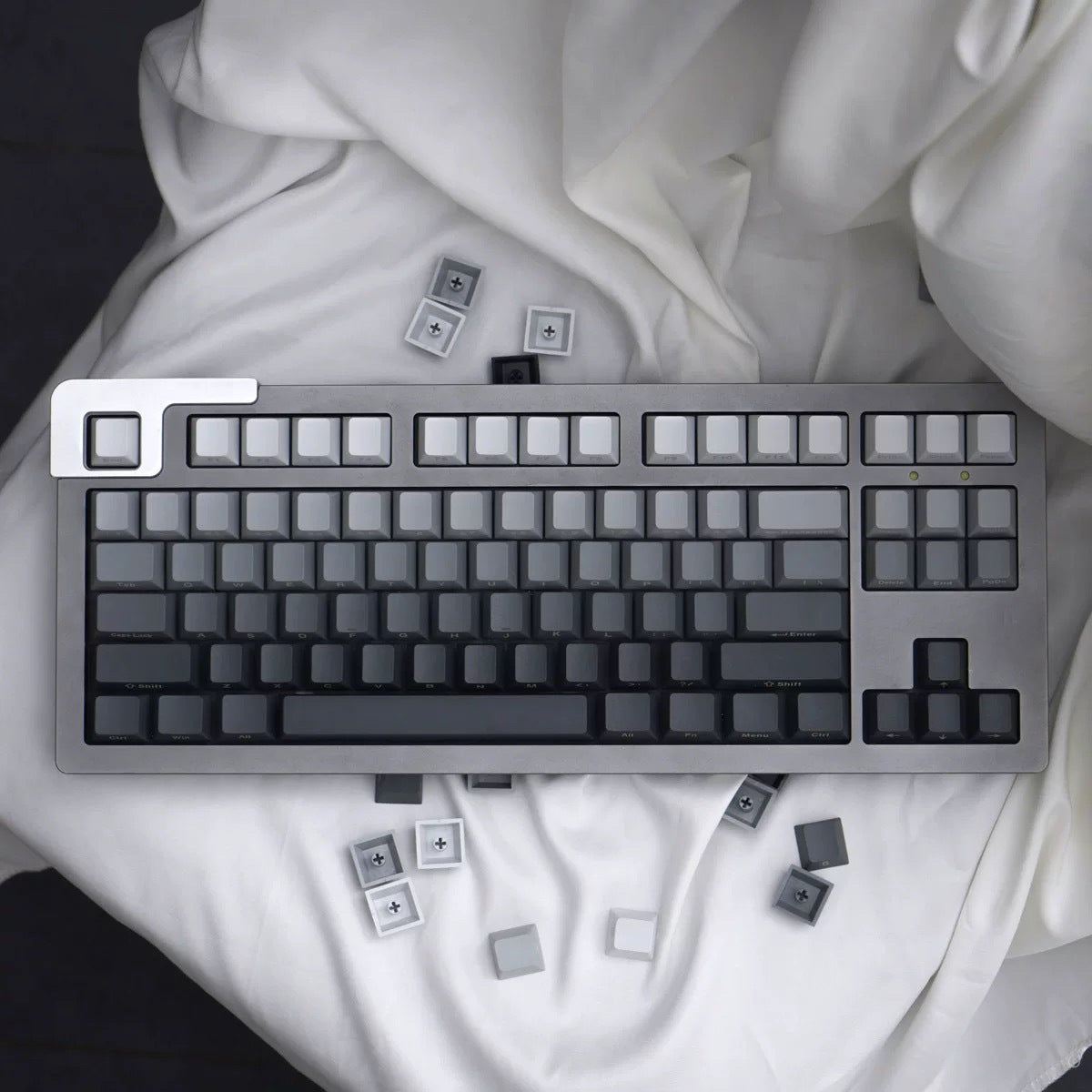 Glasskey – Top-down view of a tenkeyless keyboard with grey gradient keycaps and loose caps scattered on white cloth