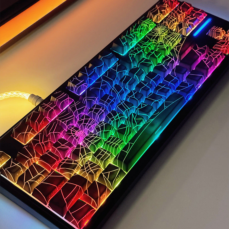 Loomwire – Full keyboard from above fitted with black spider web keycaps, backlit in a rainbow gradient