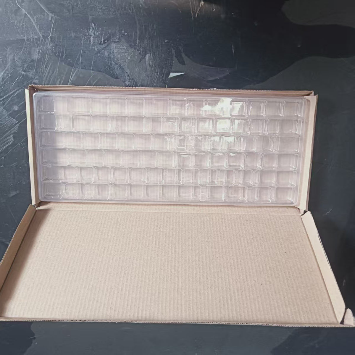 Claripack – Clear keycap tray with grid of compartments inside an open cardboard shipping box on a dark surface