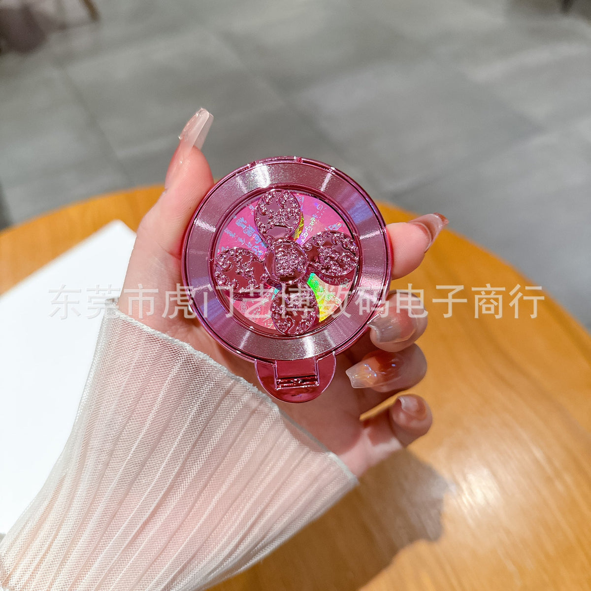 Swivelnest Rotating Phone Stand in pink metallic finish with clover spinner, held in a hand over a wooden table