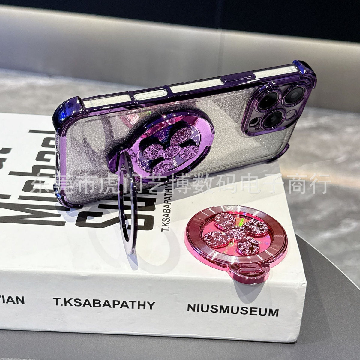 Swivelnest – Purple glitter phone case with ring stand propped on a book, a pink clover-spinner stand lying beside it