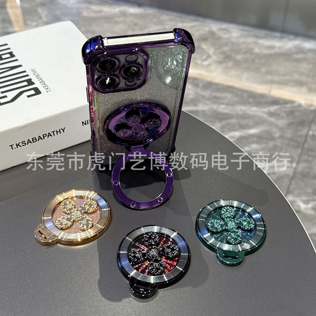 Swivelnest – Gold, black and green clover-spinner ring stands on a table beside a phone with the purple stand open