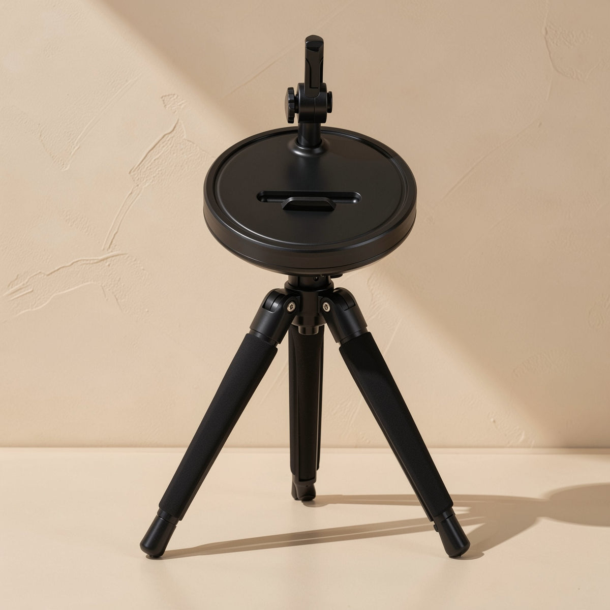 Hoverhold Overhead Phone Stand – black round base with a slot, folded tripod legs and a phone clamp arm on a beige background