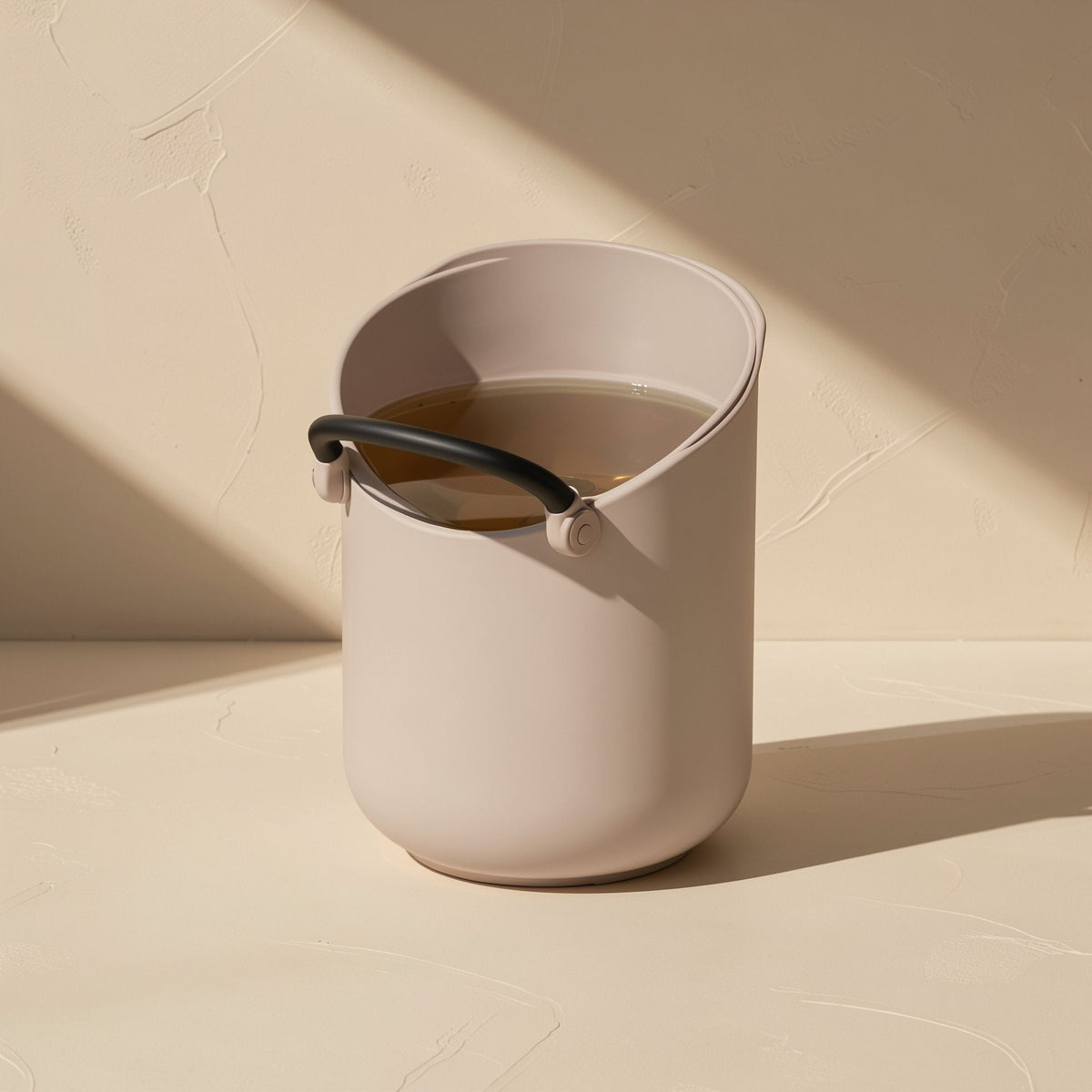 Cliphear – Taupe bucket-shaped container with a black handle holding brown liquid, on a beige surface in angled light