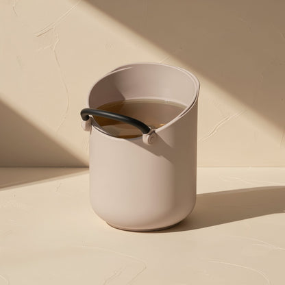 Cliphear – Taupe bucket-shaped container with a black handle holding brown liquid, on a beige surface in angled light
