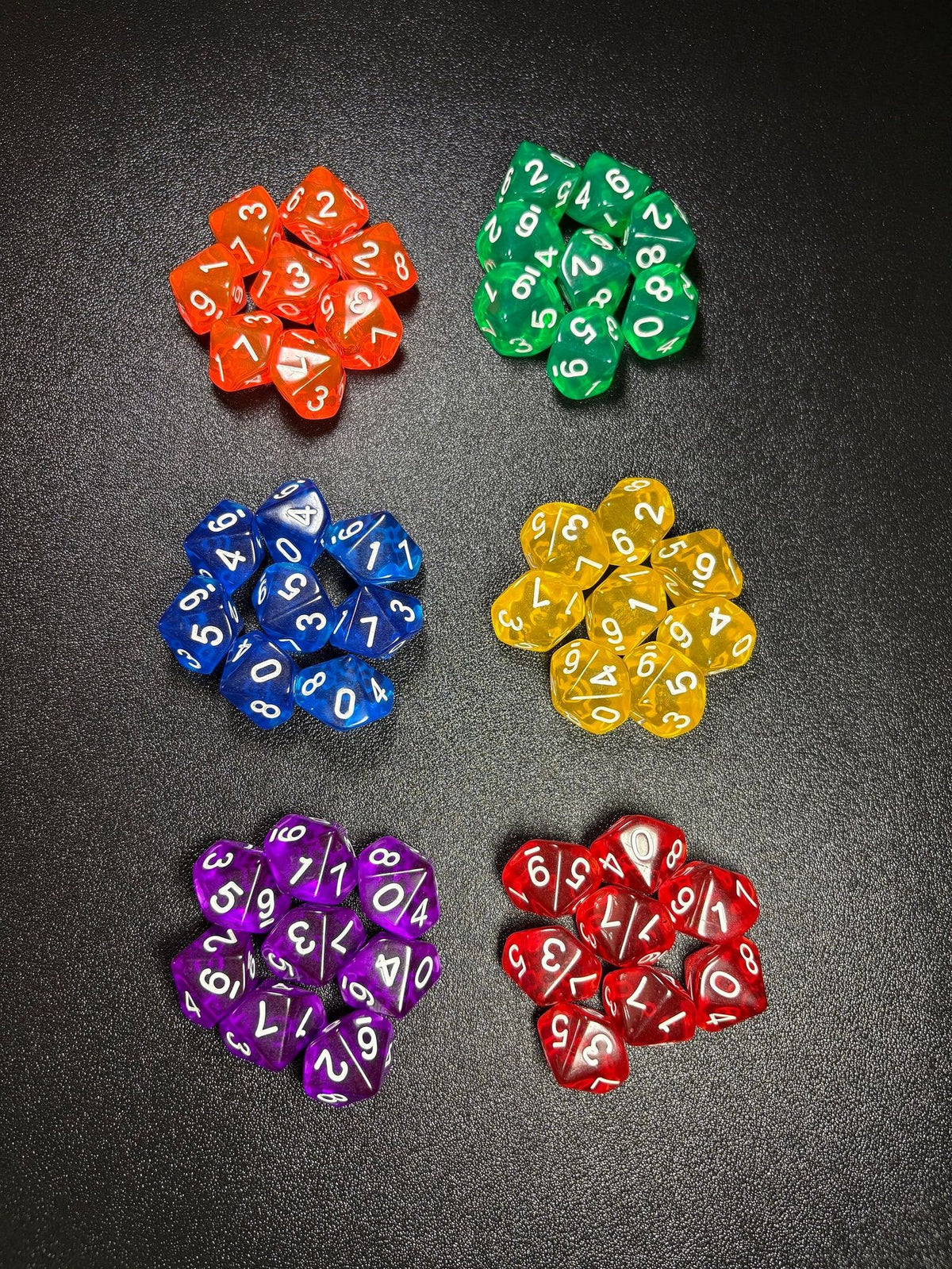 Prismdot ten-sided transparent acrylic dice set in six colors, sorted into piles on a dark surface