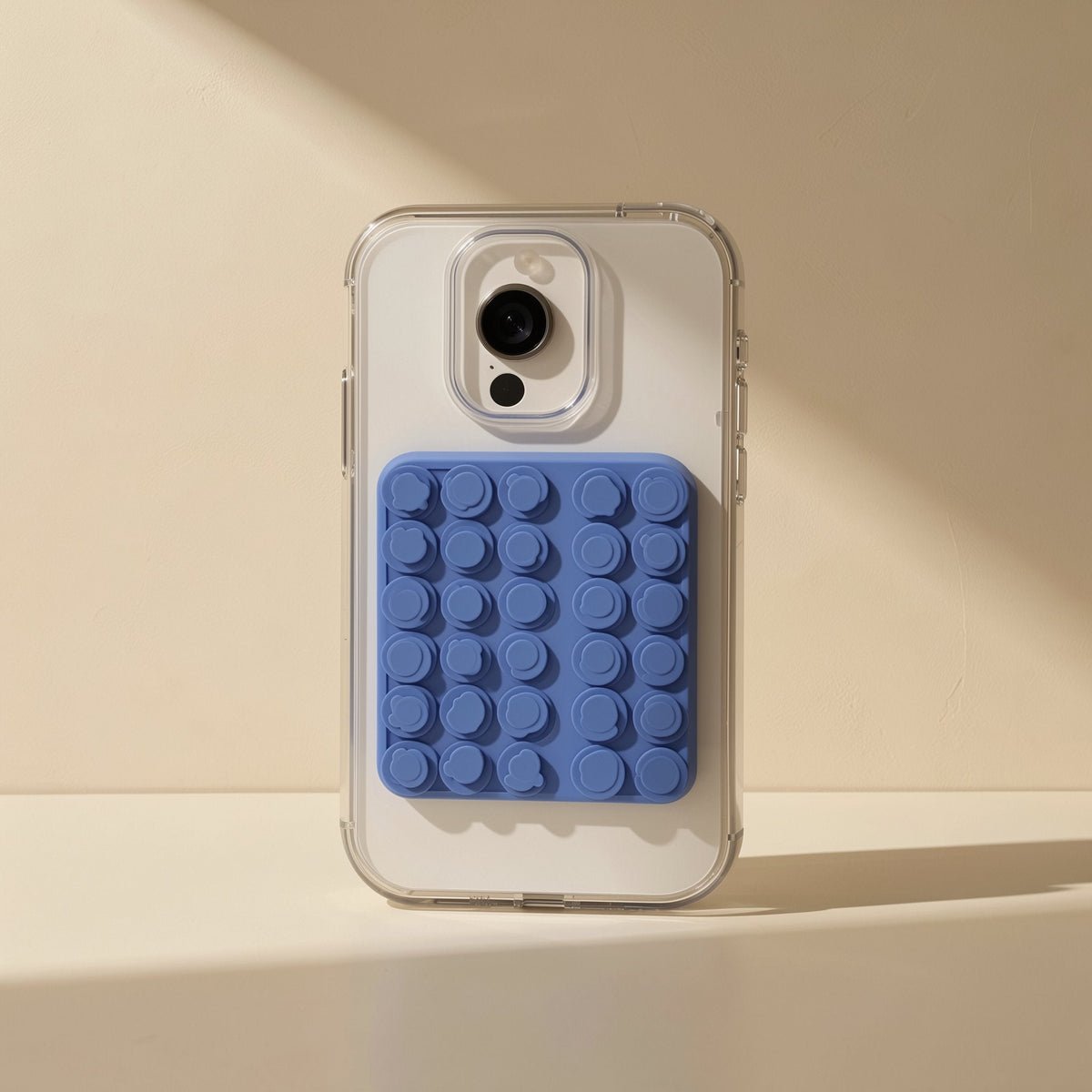 Gripcup Silicone Suction Cup Phone Stand in blue stuck to the back of a clear phone case, sunlit beige backdrop